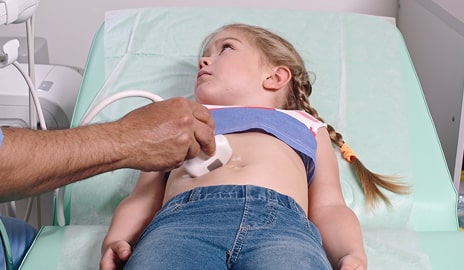 A child lies on an examination table receiving an ultrasound in a hospital room, with medical staff present.